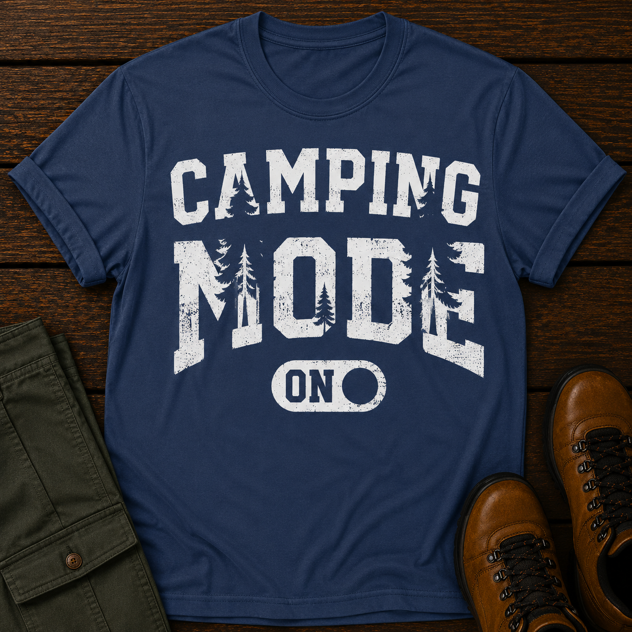 Blue t-shirt with 'Camping Mode On' text on a wooden surface with hiking pants and boots.