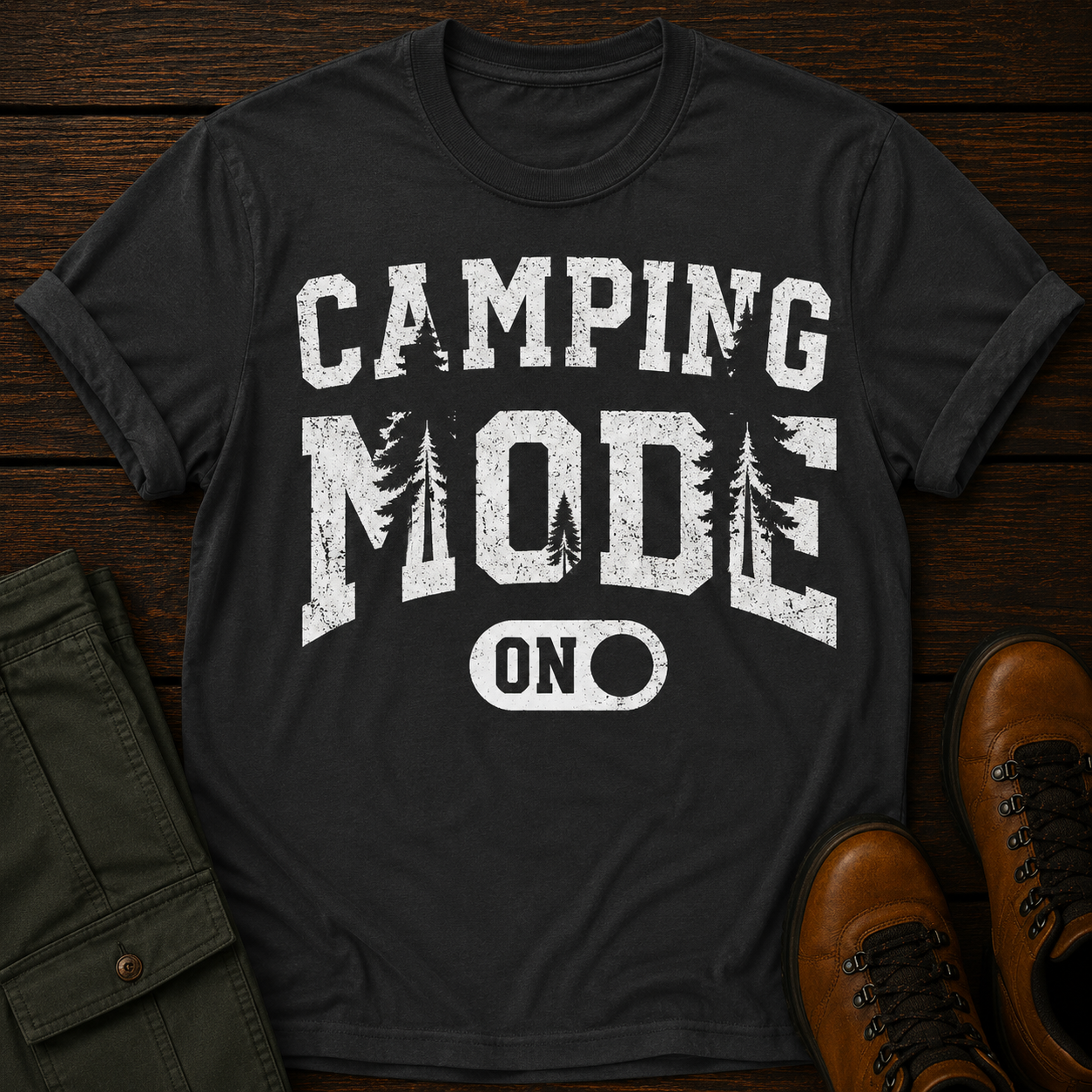 Black t-shirt with 'Camping Mode ON' text on a wooden surface with a jacket and boots.