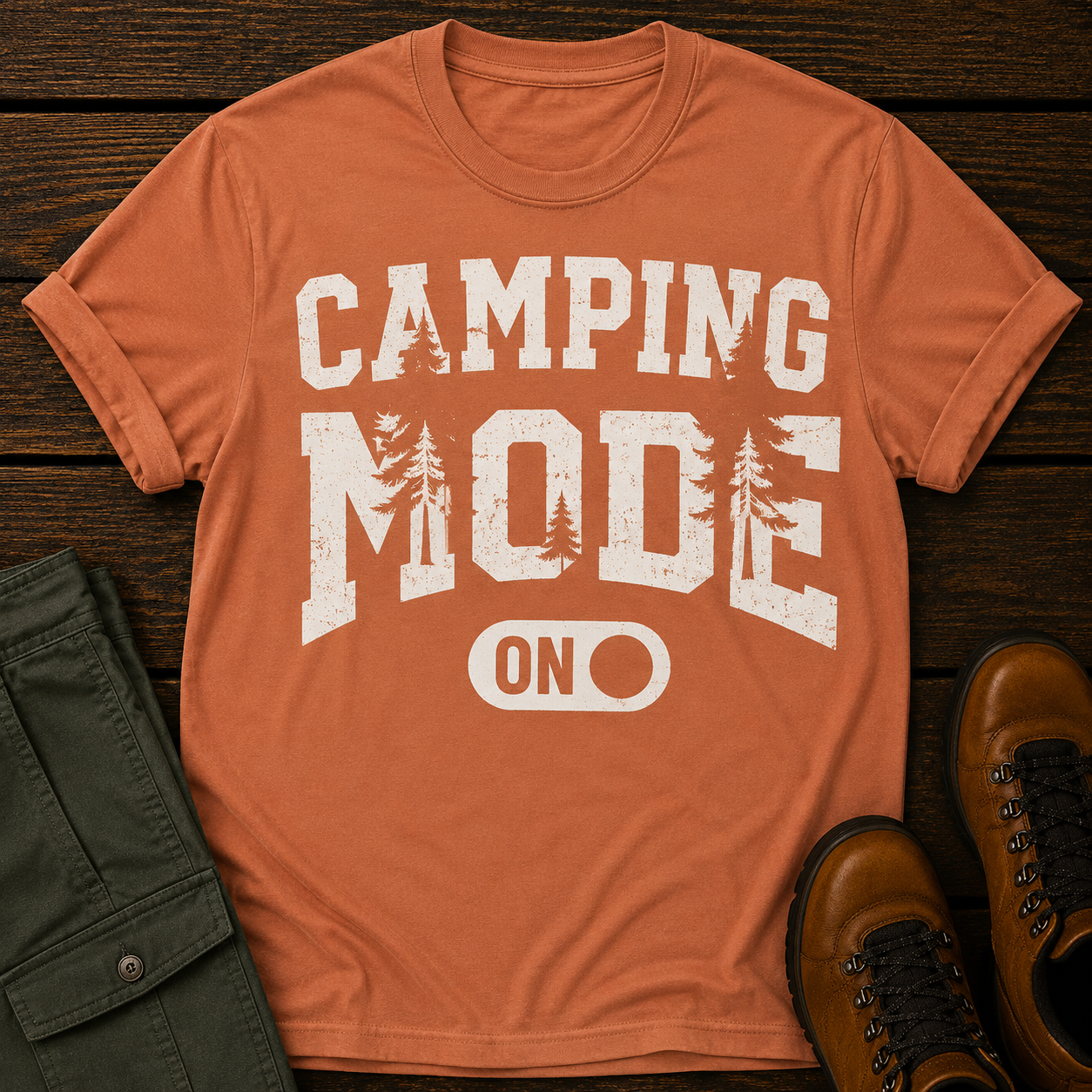 Orange t-shirt with 'Camping Mode ON' text on a wooden surface with pants and boots.