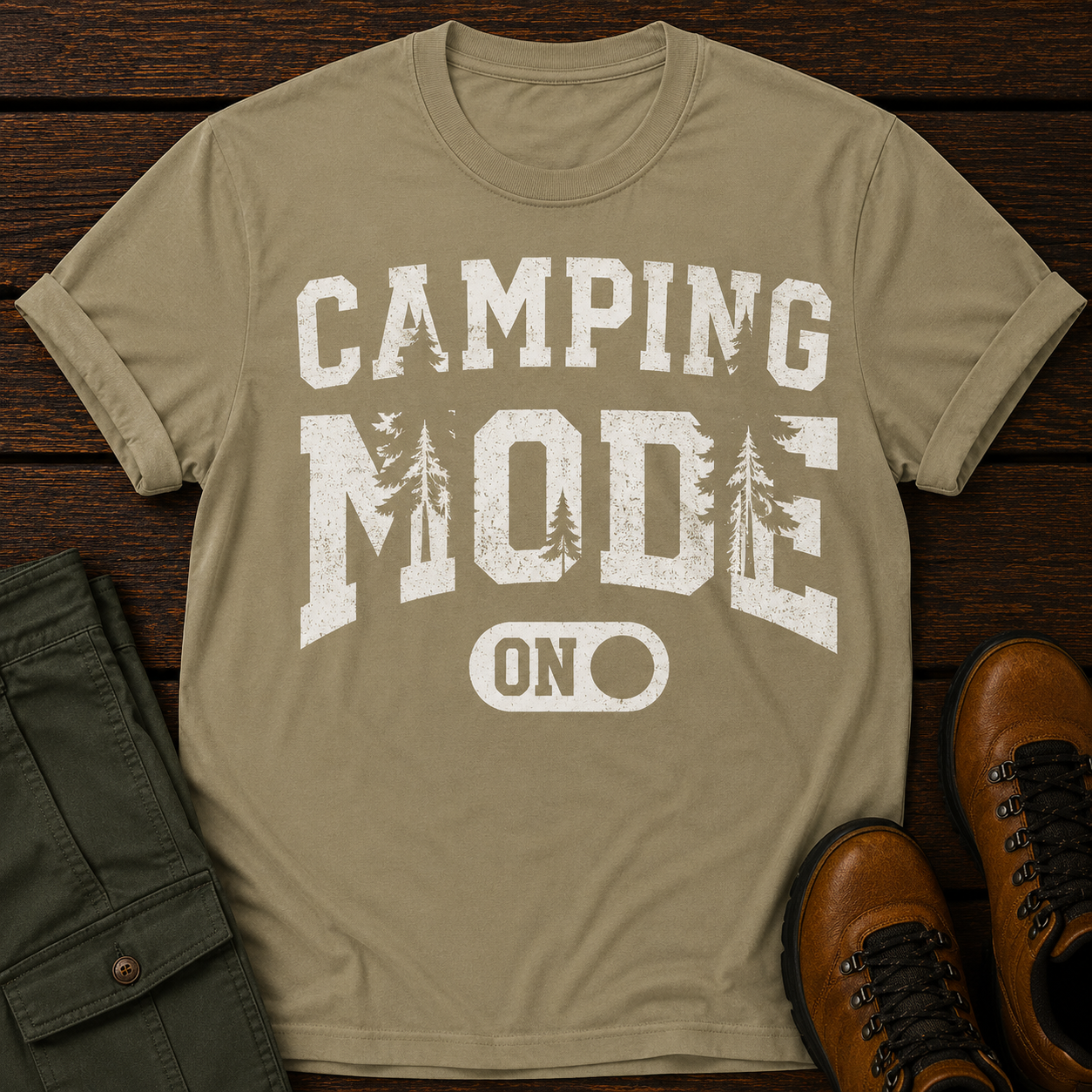 Khaki t-shirt with 'Camping Mode On' text on a wooden surface with green pants and brown boots.