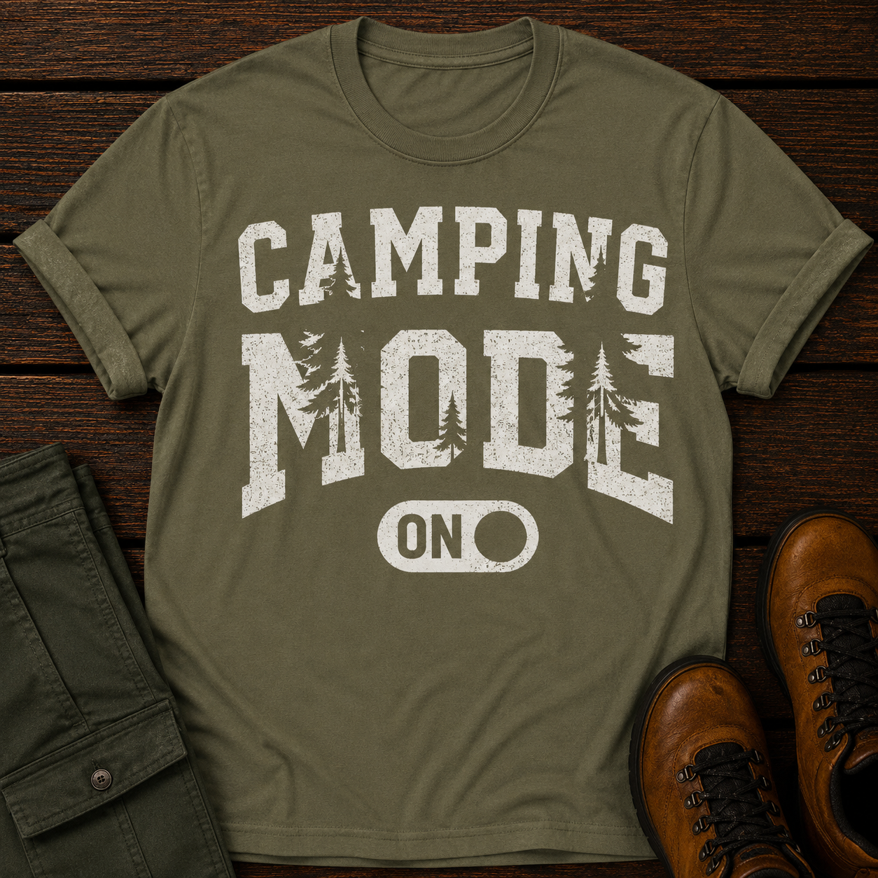Green t-shirt with 'Camping Mode On' text on a wooden surface with green pants and brown boots.