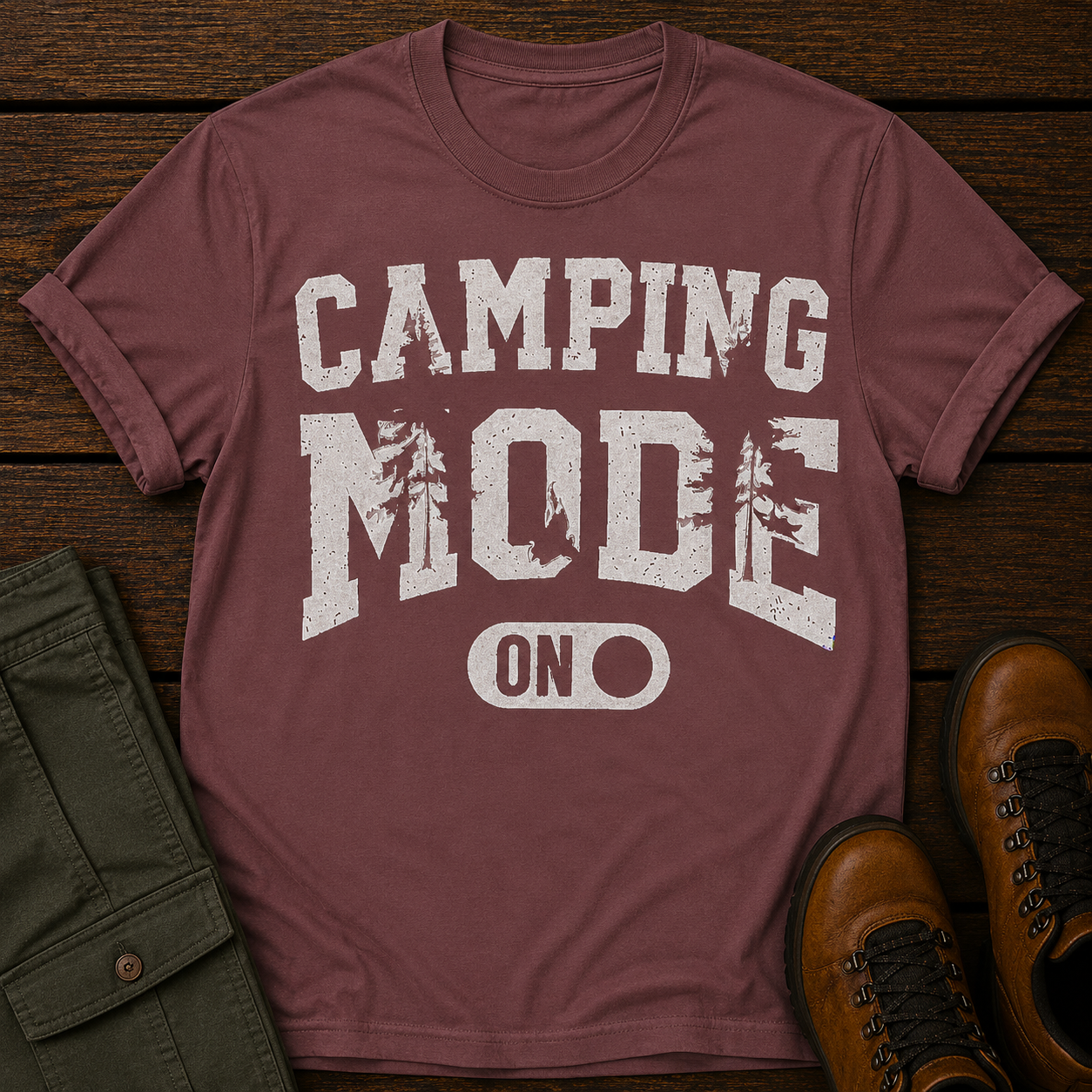 Maroon t-shirt with 'Camping Mode ON' text on a wooden surface with green pants and brown boots.